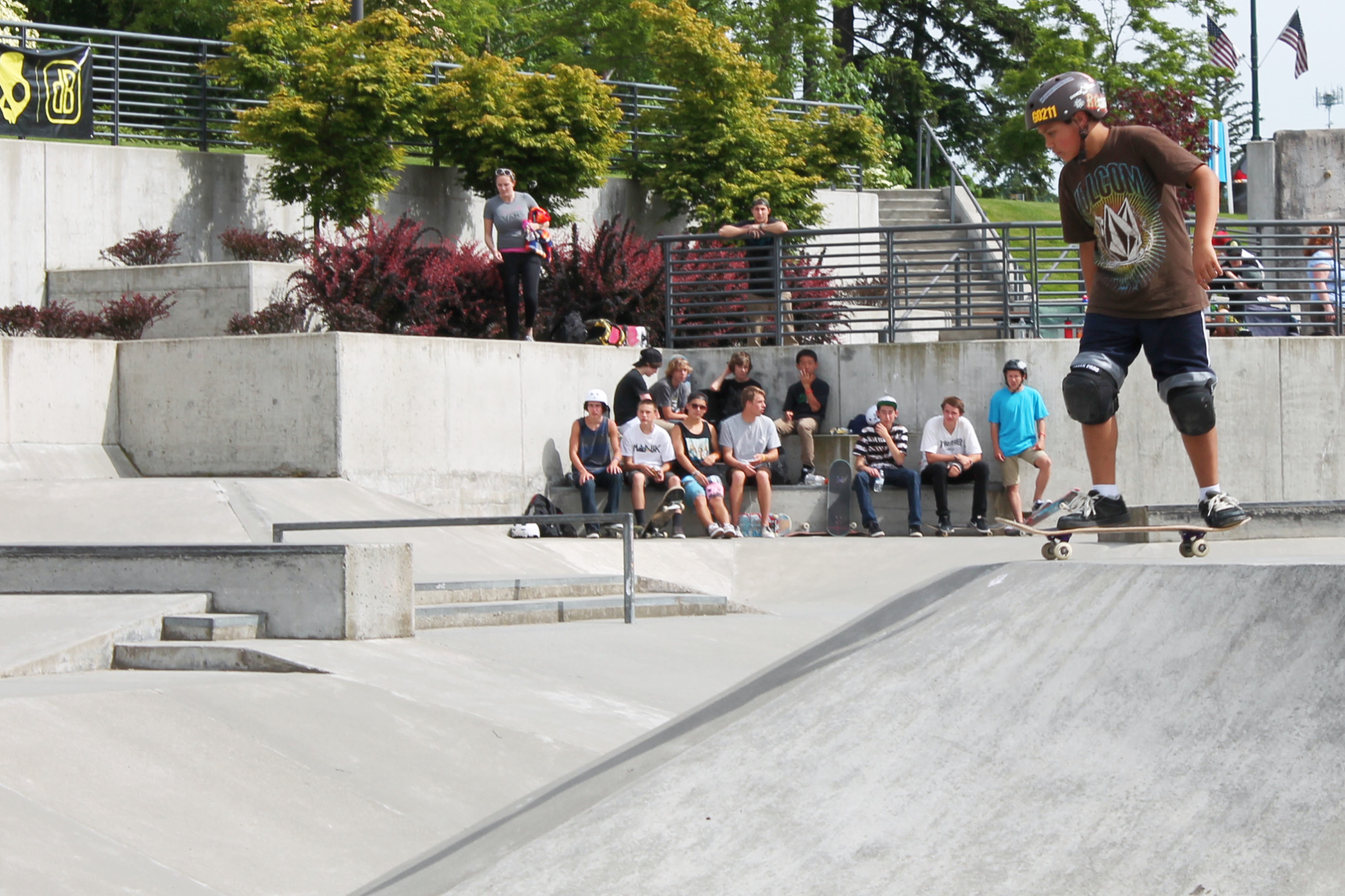 Kid in safety gear stands on skateboard at the edge of a ramp at the Sammamish Commons Skate Park while onlookers watch from the built-in cement seats.