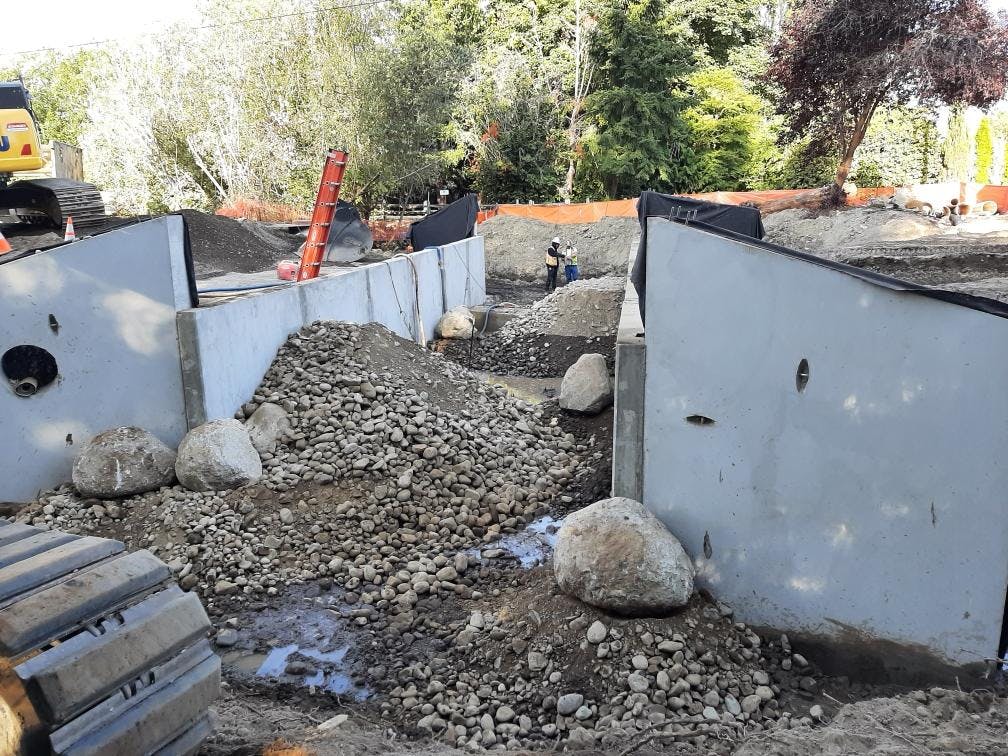 Inside the culvert during construction on the Ebright Creek Fish Passage Project. The walls are up and there are large piles of dirt and rock with a few huge boulders. Two crew members are in the background.