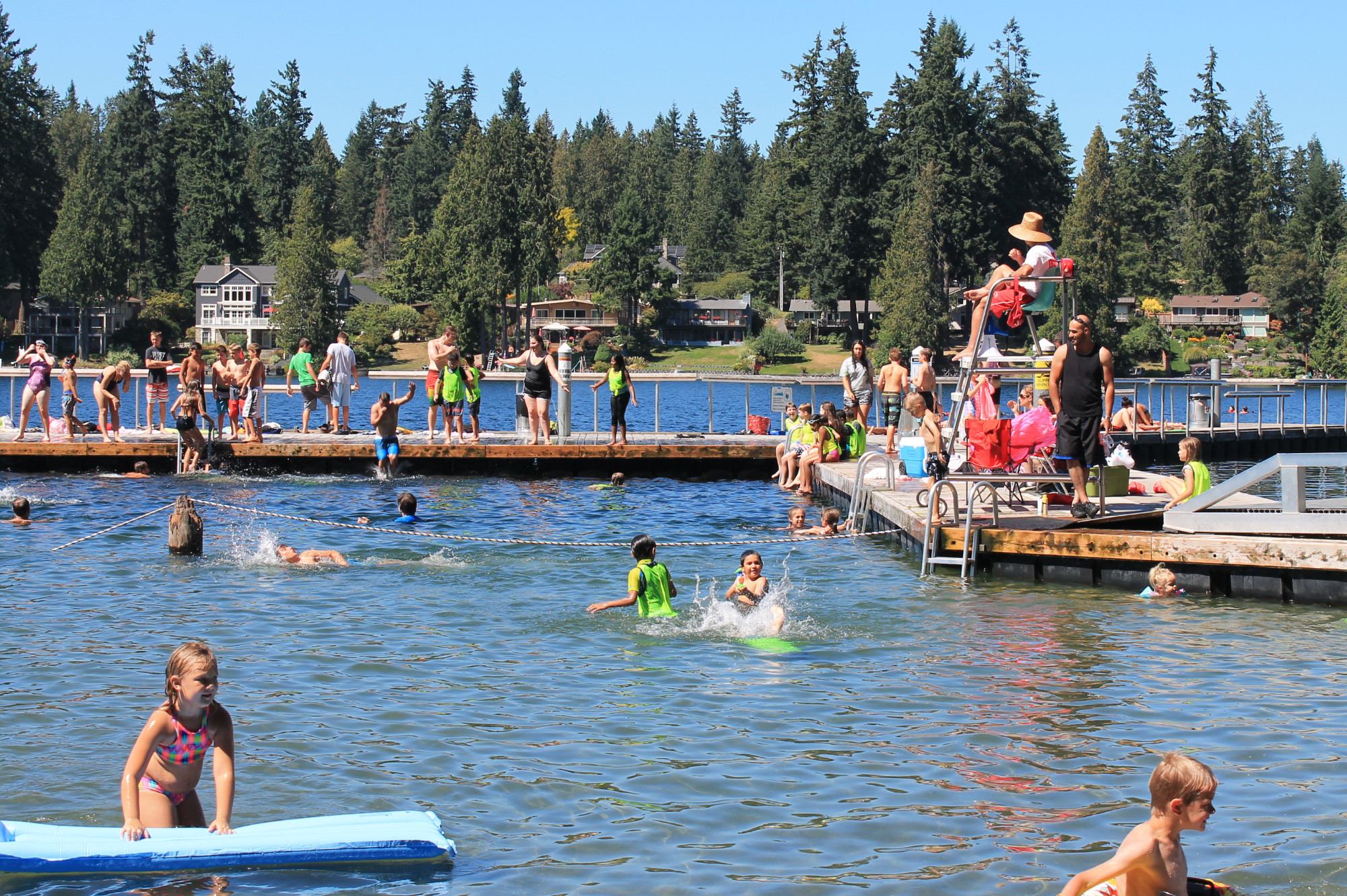 busy day at Pine Lake Park, with lots of kids wading in the shallow area and people swimming while a lifeguard watches from the dock