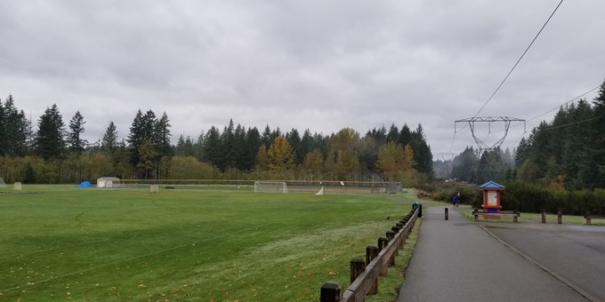 walkway beside a football field, suspension lines to the side, a map kiosk available, and a conifer forest in the distance