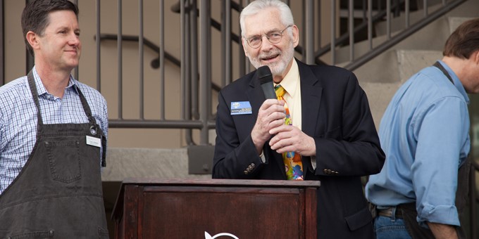 person in a suit with colorful tie smiles while speaking into a microphone at a Metropolitan Market branded podium, an aproned worker beside