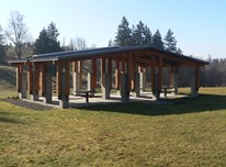 Large covered picnic shelter at the Lower Commons at Sammamish Commons with several picnic tables. The floor is cement. The shelter is surrounded by grass and there are evergreen trees and deciduous trees without their leaves in the background.