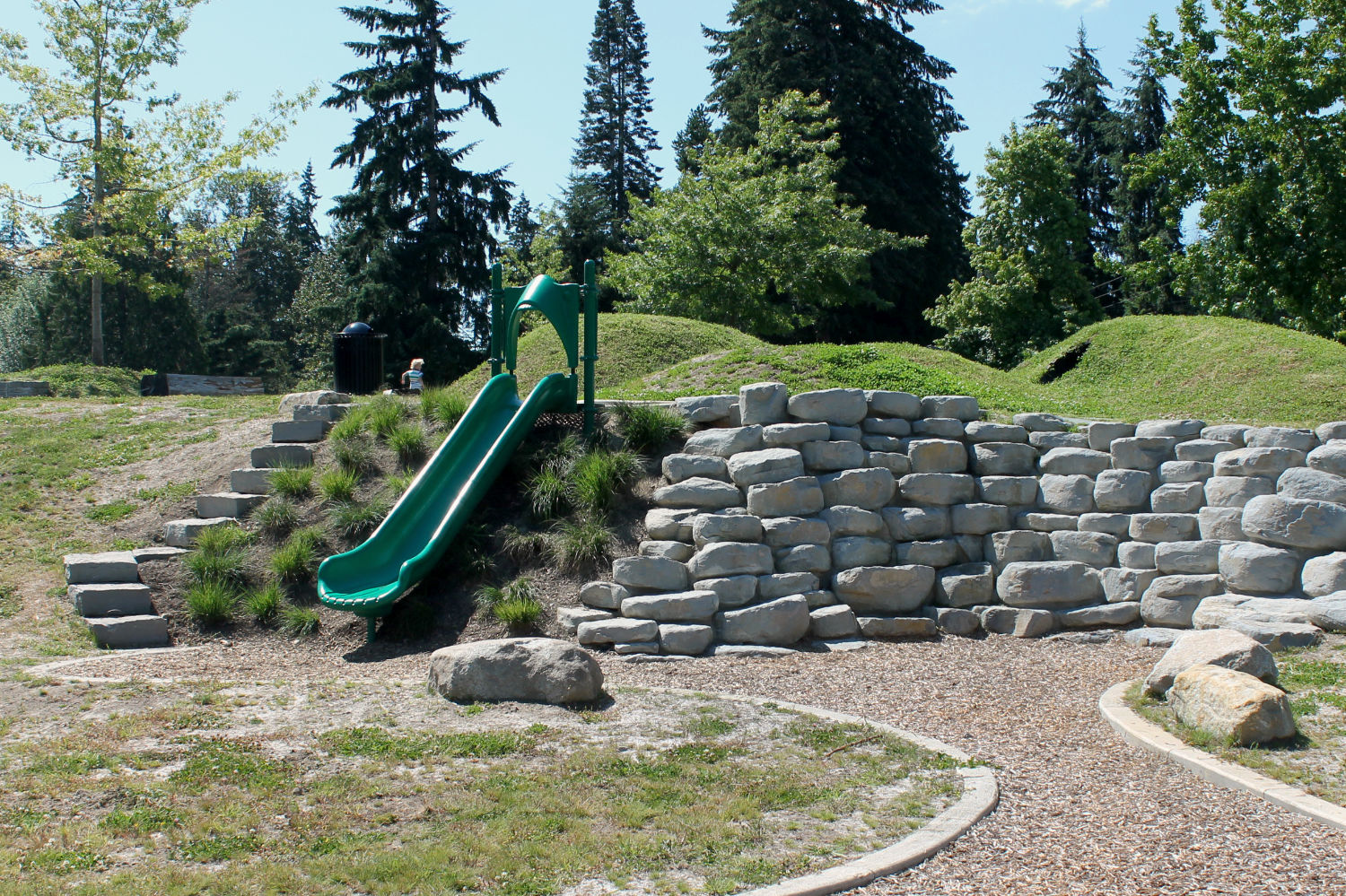 short slide beside a rock wall at big rock park north, with small grass hillocks at the top of the slide and stairs to access