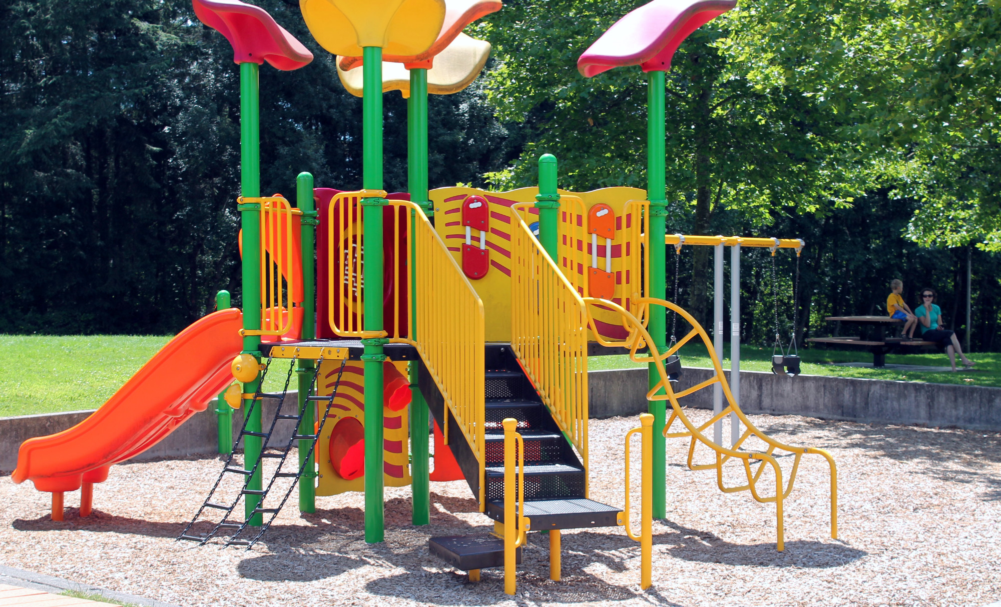 Colorful play structure containing a slide, climbing bars and ropes, and swings. Wood chips cover the play area. An adult and child sit at a picnic table behind.