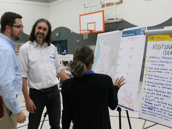 Three people stand in front of an informational poster board on a tripod while having a discussion during a public workshop regarding the Transportation Master Plan.