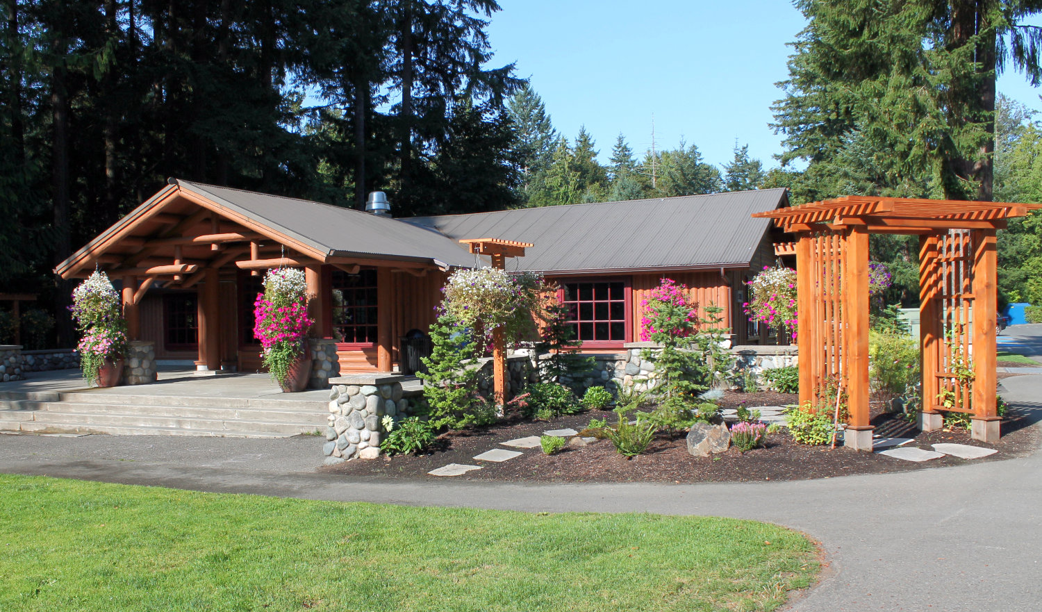 Exterior of Beaver Lake Lodge showing front entrance lined by welcoming giant vases and hanging baskets full of flowers. Small landscaped area in front has small plants, stepping stones, and a wooden arbor.