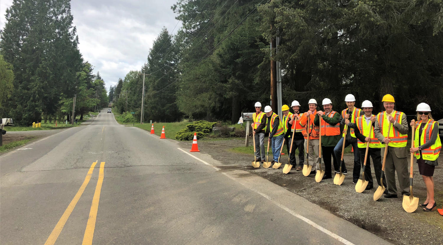 ten officials and project members with golden shovels celebrate groundbreaking on the gravel shoulder of the existing road
