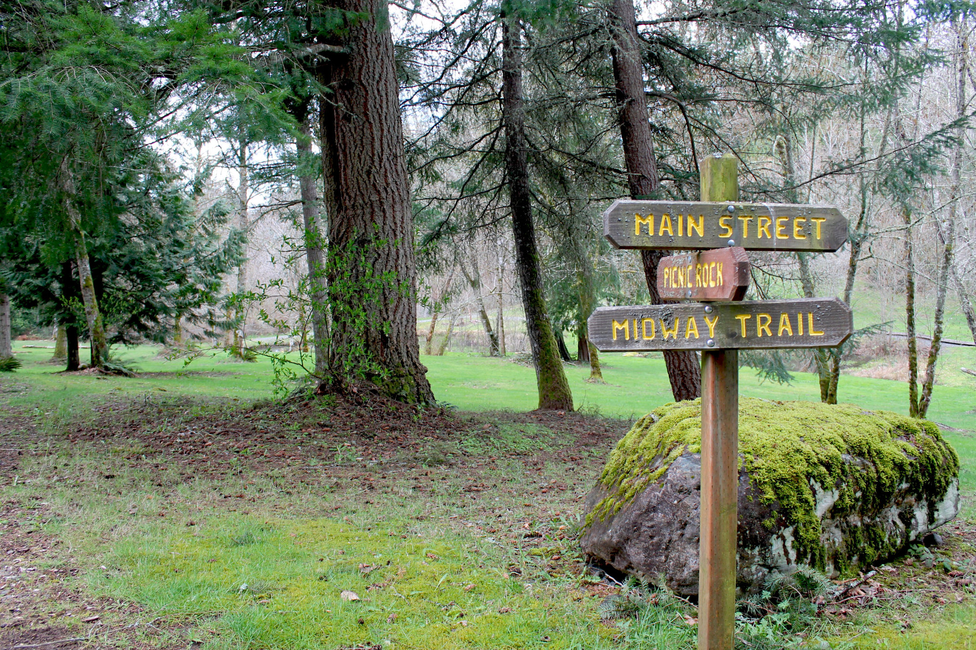 Sign at Big Rock Park trail intersection points the different ways to Main Street, Midway Trail, and Picnic Rock. A giant moss-covered rock sits next to the sign. There are trees and a grassy area in the background.