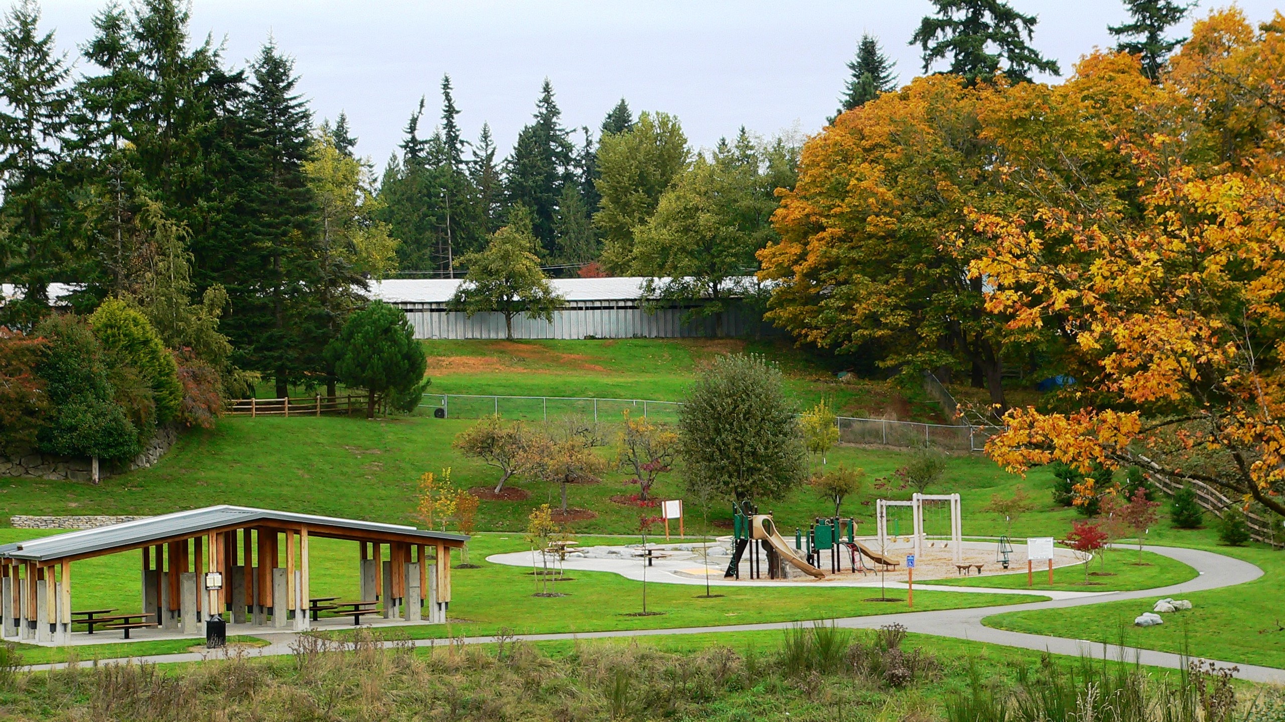 Covered picnic shelter with multiple picnic tables and children's playground with slides, swings, bars, and a variety of other playground equipment, surrounded by grassy areas which are then bordered by deciduous and coniferous trees. 