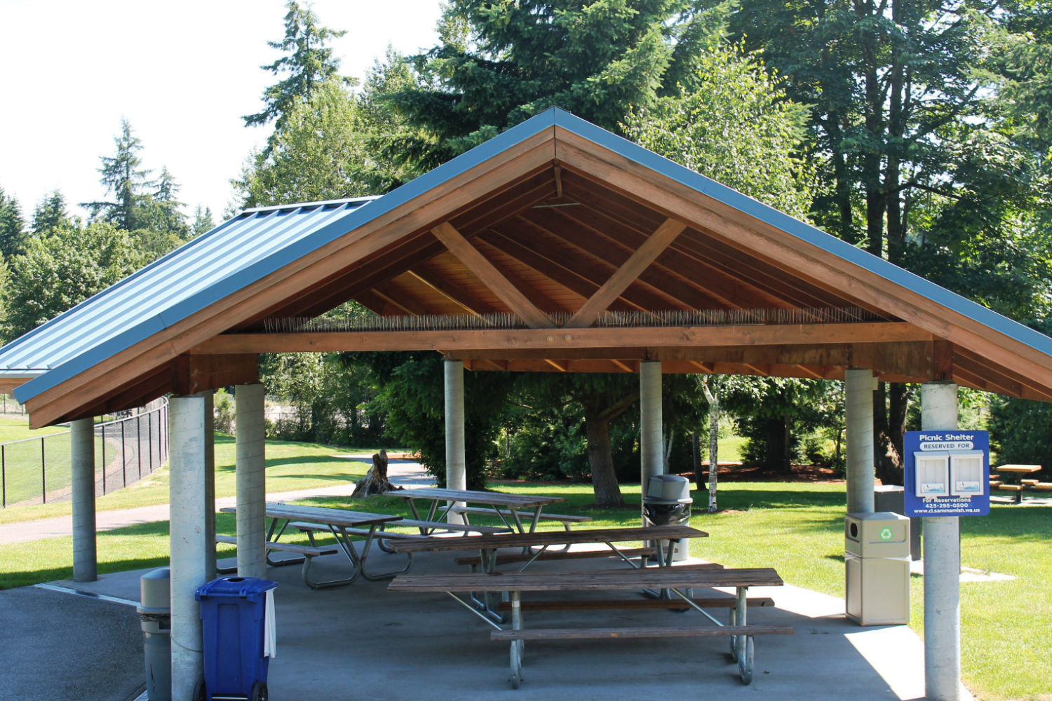 East Sammamish Park picnic shelter with blue metal roof and four picnic tables. There are four garbage/recycle bins. The shelter is surrounded by a grassy area, trees, and near a fenced field.