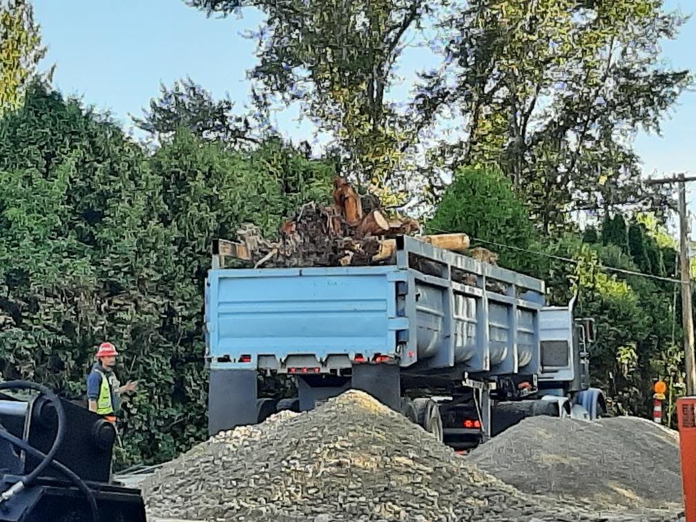 A crew member working on the Ebright Creek Fish Passage Project stands next to piles of rock and soil and a truck full of wood debris.