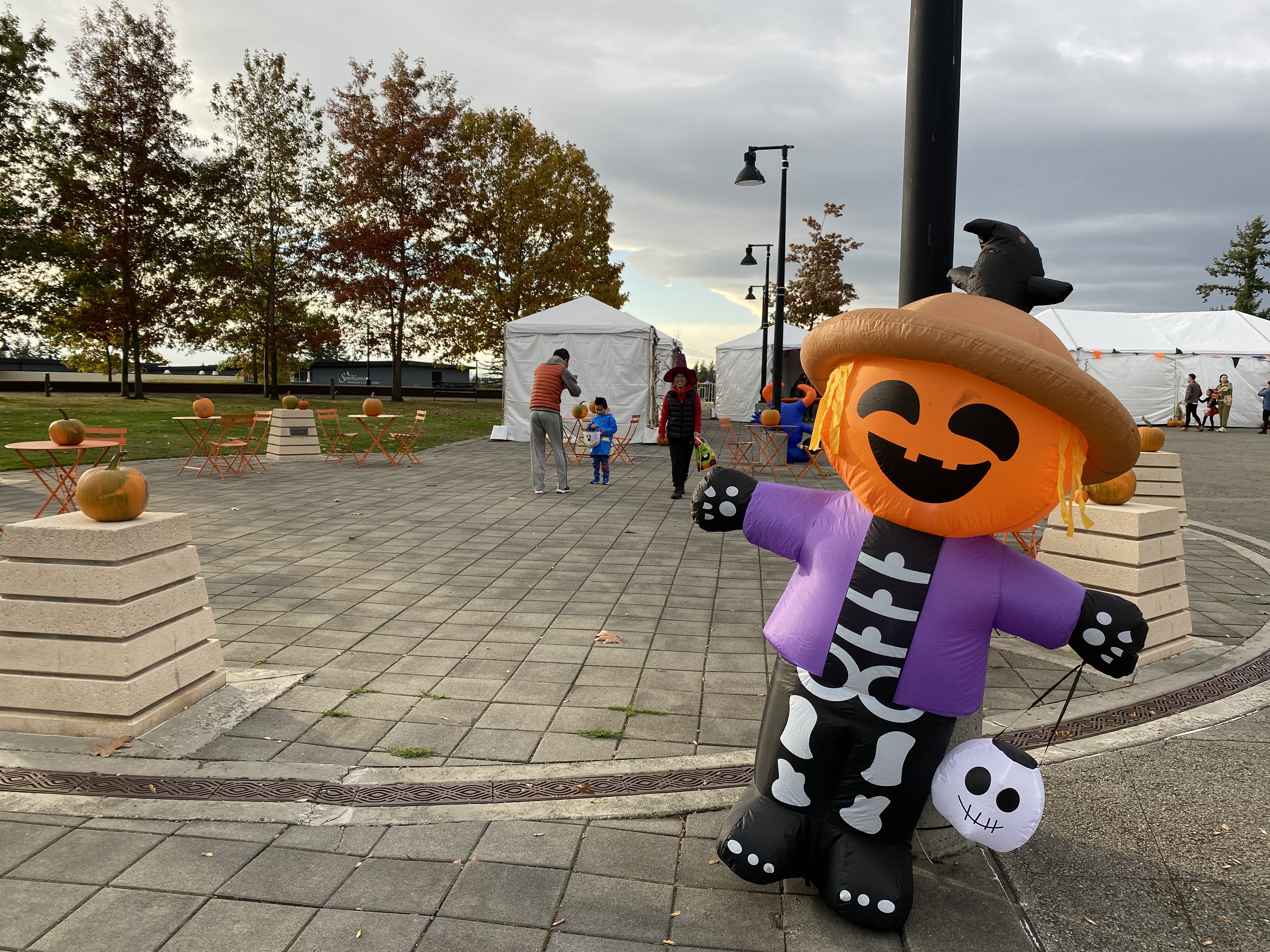 Pumpkins and other Halloween decorations adorn the Sammamish Plaza. White event tents cover booths, and trick-or-treating families walk in the background. 