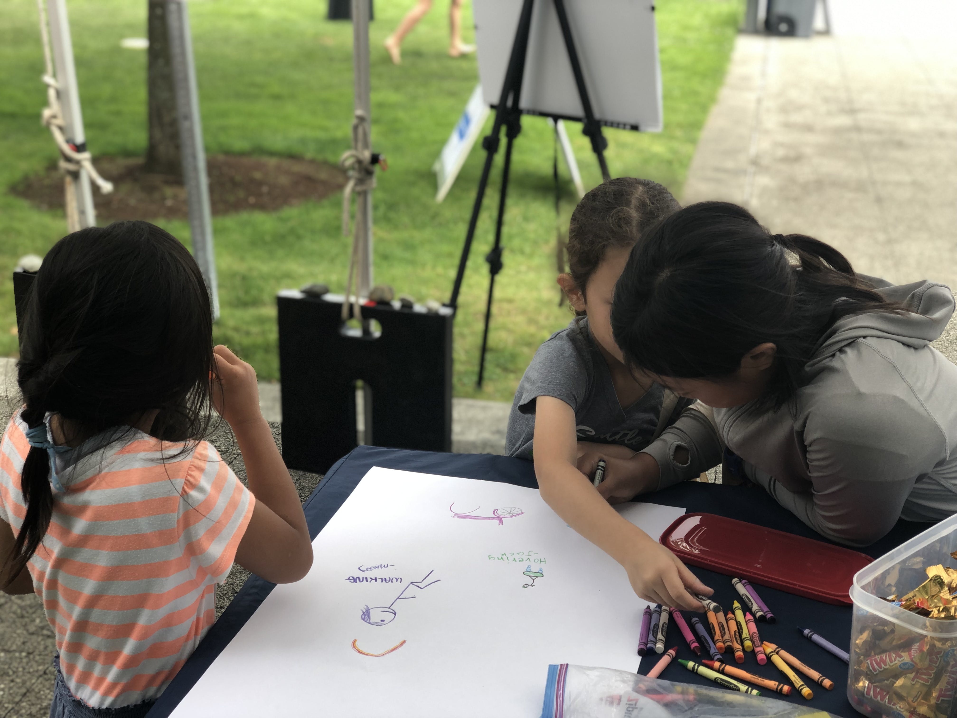 Three children use crayons to draw different types of transportation on a shared piece of paper at Party on the Plateau.