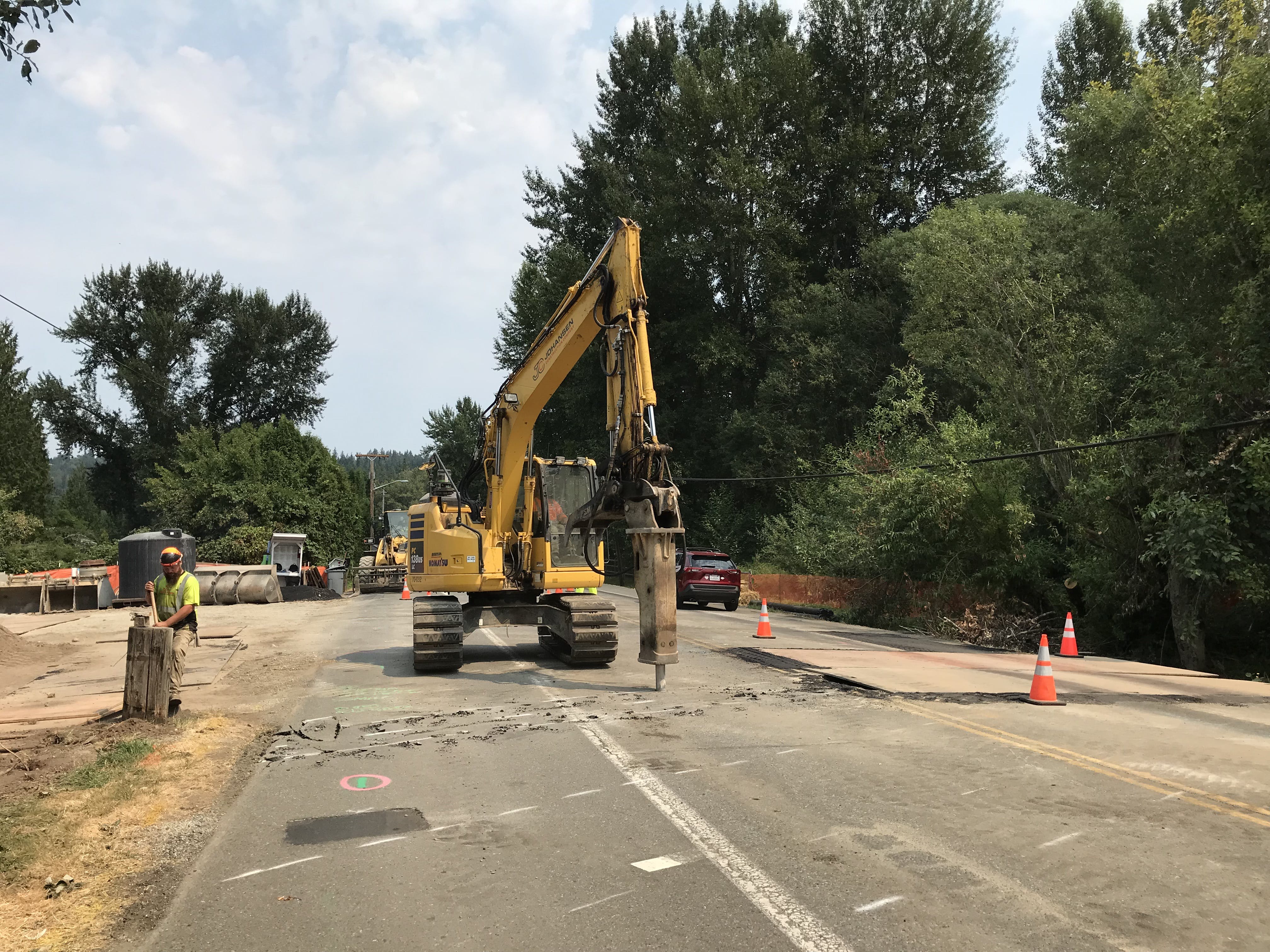 Yellow excavator with a hydraulic breaker, preparing to install the dewatering system for the Ebright Creek Fish Passage project in Sammamish while crew member works on the road side.