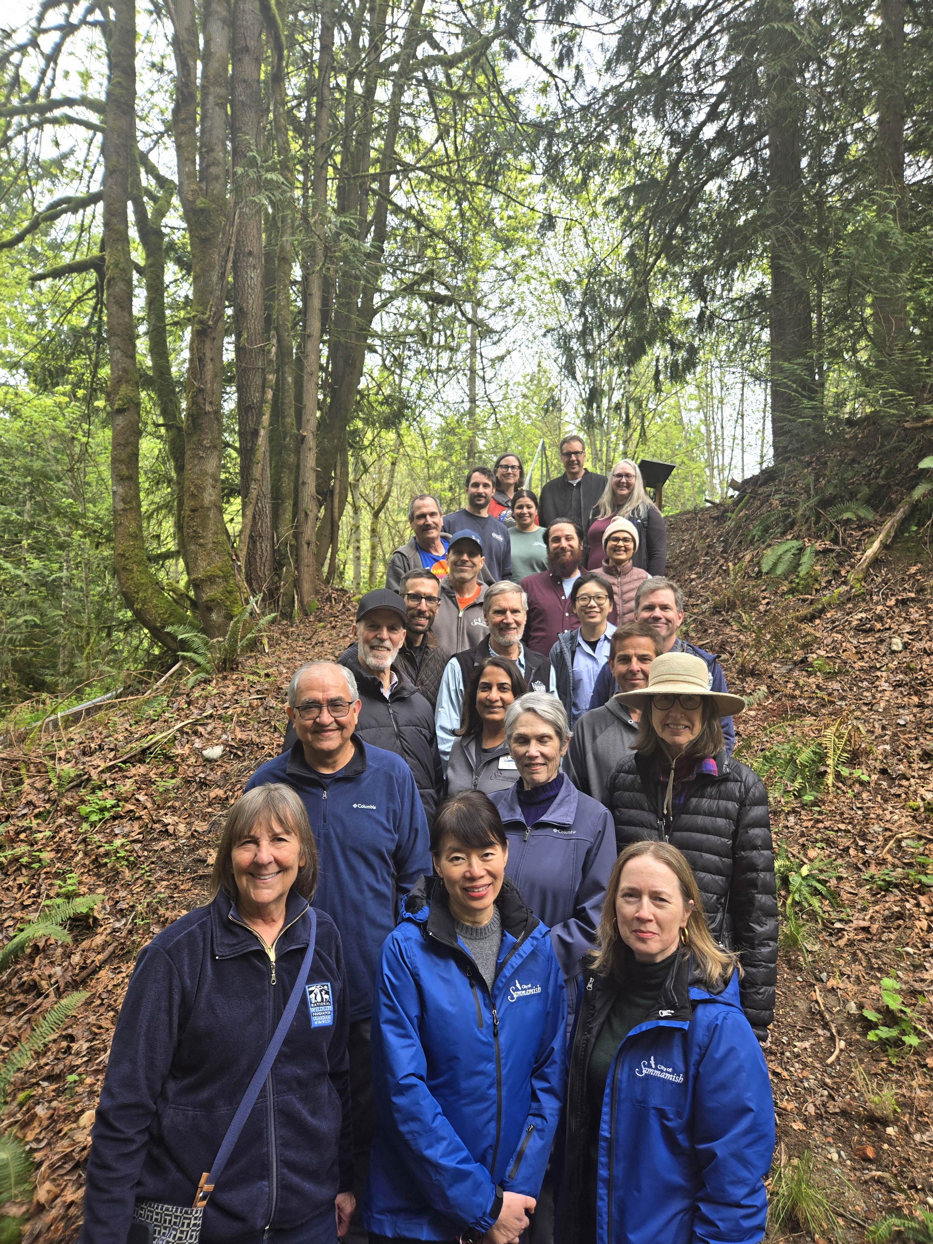 A diverse group of 24 adults smiling in a forest setting, wearing jackets and casual outdoor attire. Tall trees and leafy ground create a serene backdrop.