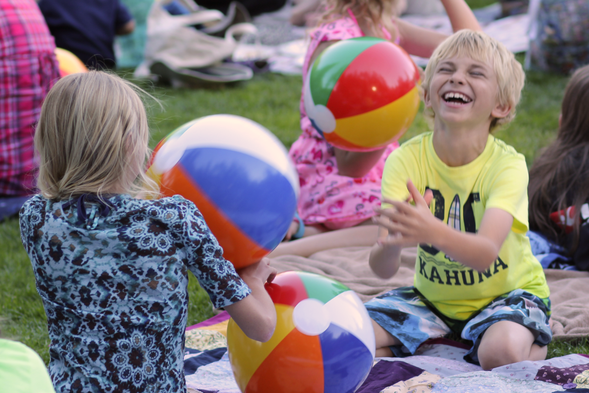 Two children laugh while playing with three beach balls during a concert in the park