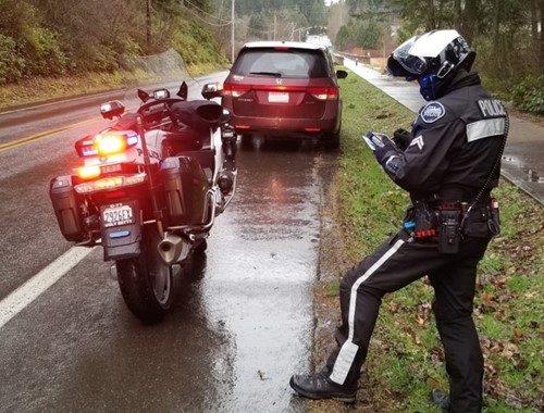 A picture of a police officer standing next to their police bike writing a ticket alongside a road where a car is stopped in front of the police bike.
