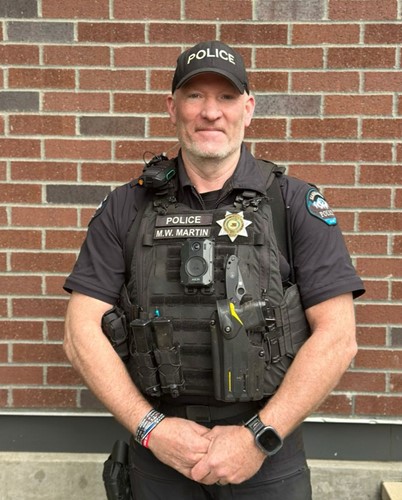 A picture of a man in a police uniform complete with a safety vest, badge, radio, and ball cap posing for a photo.