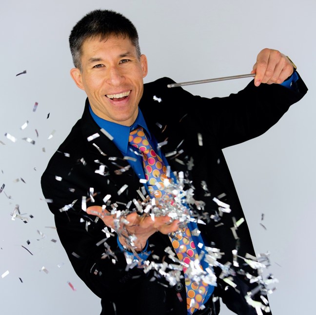 Magician Jeff Evans holds a magic wand in one hand and an explosion of metallic confetti erupts from his other hand.