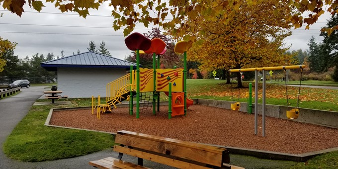 Children's playground unit containing a slide and climbing features and a separate swing set are featured in the Klahanie Park Playground. A park bench sits in the foreground and a picnic table and restroom facilities are in the background. Golden autumn leaves cover the grassy area behind the playground and are in the deciduous trees nearby.