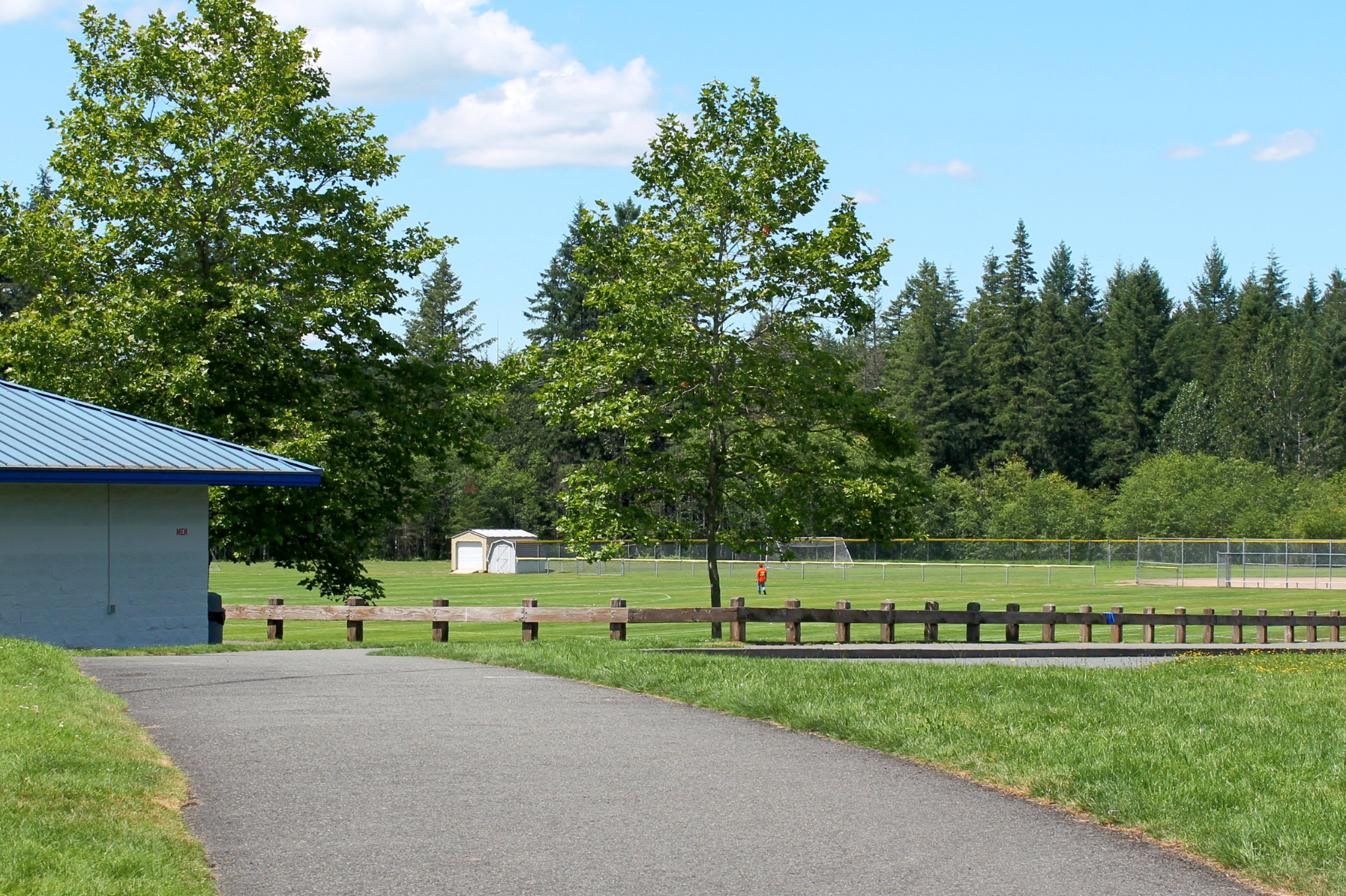 Paved path at Klahanie Park leading to the restrooms and overlooking the sports field. A railing separates the path from the fields.