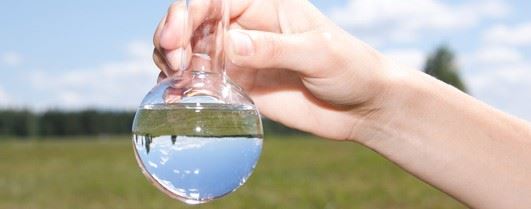 Close up shot of a hand holding a water-filled glass round laboratory flask outdoors as if a water sample was just collected.