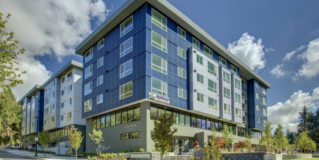 rendering of SAMM Apartments, showing a block of several multi-story buildings in white, blue, and gray, with a raised patio or plaza at one end